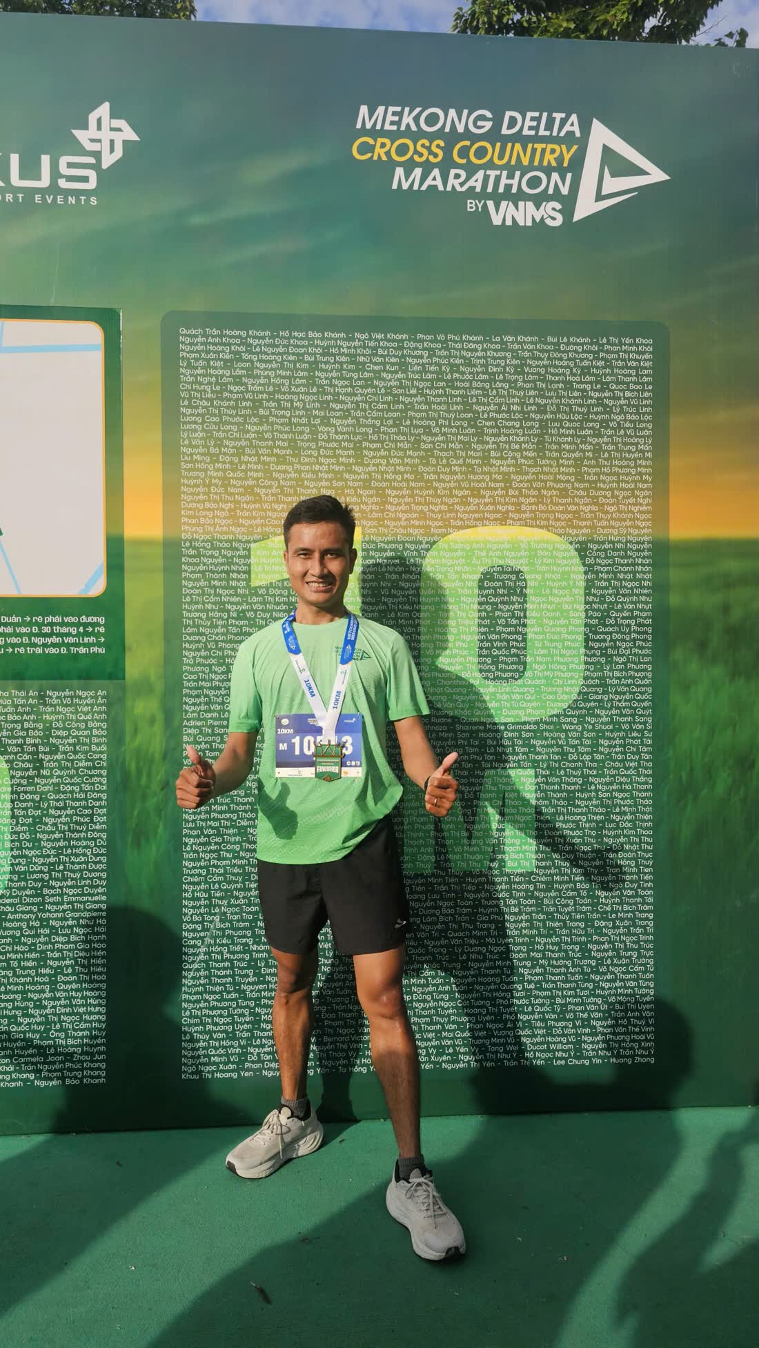 Minh mặc áo xanh, đeo huy chương, giơ hai tay trước bảng giải Mekong Delta Cross Country Marathon